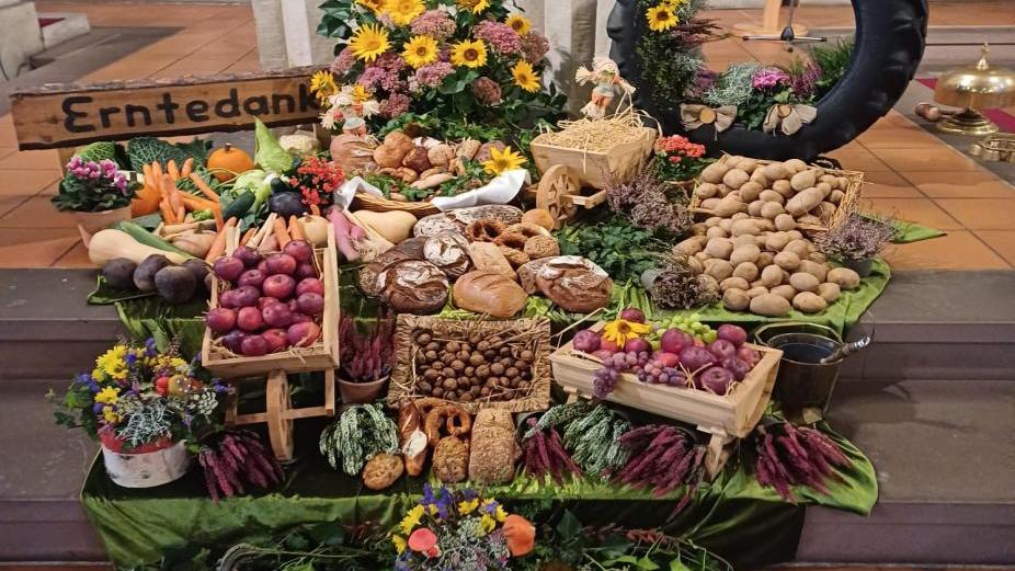 Auf dem Bild ist ein reich geschmückter Erntedankaltar in einer Kirche zu sehen. Auf mehreren Ebenen sind frische Erntegaben arrangiert: Kartoffeln, Karotten, Rote Bete, Äpfel, Trauben, Brotlaibe, Nüsse, Kürbisse, Zwiebeln und weitere Gemüse- und Obstsorten. Bunte Blumen, grüne Tücher und dekorative Elemente wie ein Reifen mit Sonnenblumen, Holzschubkarren und ein „Erntedank“-Schild aus Holz runden das festliche Arrangement ab. Im Hintergrund steht ein mit weißer Altardecke geschmückter Altar.