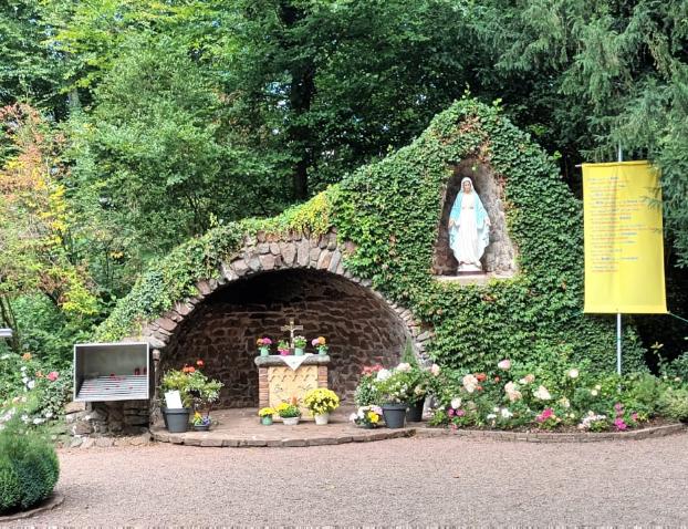 „Mariengrotte im Grünen, überwuchert von Efeu und Blumen. In der Mitte befindet sich ein kleiner Altar mit Blumenschmuck, darüber in einer Nische eine Statue der Jungfrau Maria. Daneben hängen Tafeln, ein großes Kreuz aus Metall sowie ein gelbes Banner mit Text. Vor der Grotte stehen Bänke und Blumengefäße