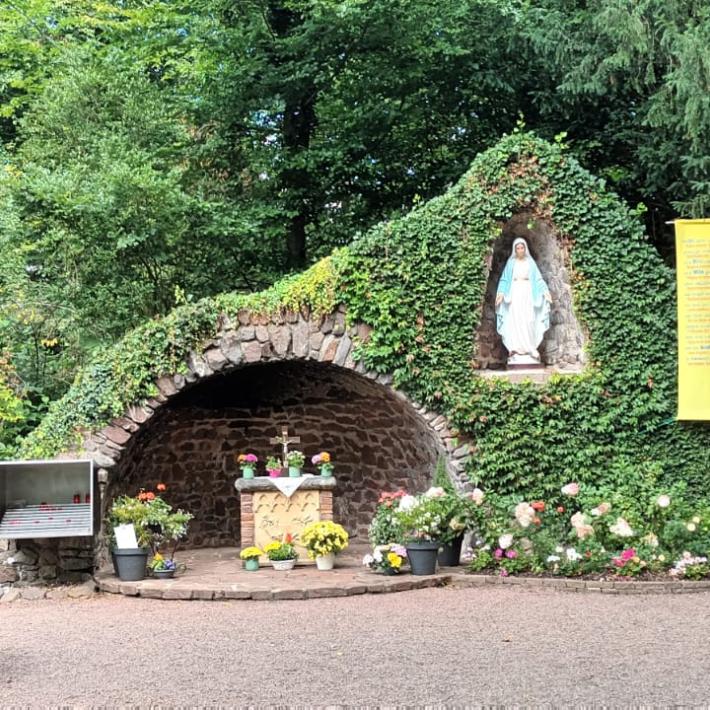 „Mariengrotte im Grünen, überwuchert von Efeu und Blumen. In der Mitte befindet sich ein kleiner Altar mit Blumenschmuck, darüber in einer Nische eine Statue der Jungfrau Maria. Daneben hängen Tafeln, ein großes Kreuz aus Metall sowie ein gelbes Banner mit Text. Vor der Grotte stehen Bänke und Blumengefäße