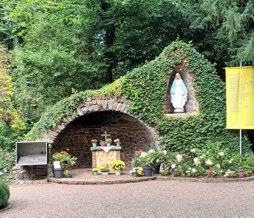 „Mariengrotte im Grünen, überwuchert von Efeu und Blumen. In der Mitte befindet sich ein kleiner Altar mit Blumenschmuck, darüber in einer Nische eine Statue der Jungfrau Maria. Daneben hängen Tafeln, ein großes Kreuz aus Metall sowie ein gelbes Banner mit Text. Vor der Grotte stehen Bänke und Blumengefäße