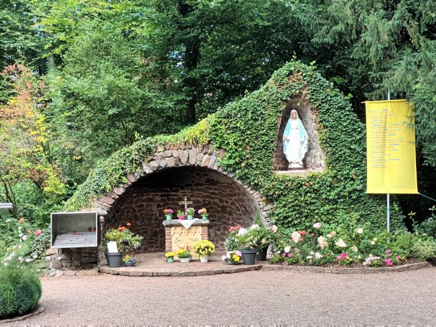 „Mariengrotte im Grünen, überwuchert von Efeu und Blumen. In der Mitte befindet sich ein kleiner Altar mit Blumenschmuck, darüber in einer Nische eine Statue der Jungfrau Maria. Daneben hängen Tafeln, ein großes Kreuz aus Metall sowie ein gelbes Banner mit Text. Vor der Grotte stehen Bänke und Blumengefäße