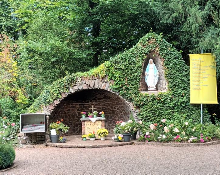 „Mariengrotte im Grünen, überwuchert von Efeu und Blumen. In der Mitte befindet sich ein kleiner Altar mit Blumenschmuck, darüber in einer Nische eine Statue der Jungfrau Maria. Daneben hängen Tafeln, ein großes Kreuz aus Metall sowie ein gelbes Banner mit Text. Vor der Grotte stehen Bänke und Blumengefäße