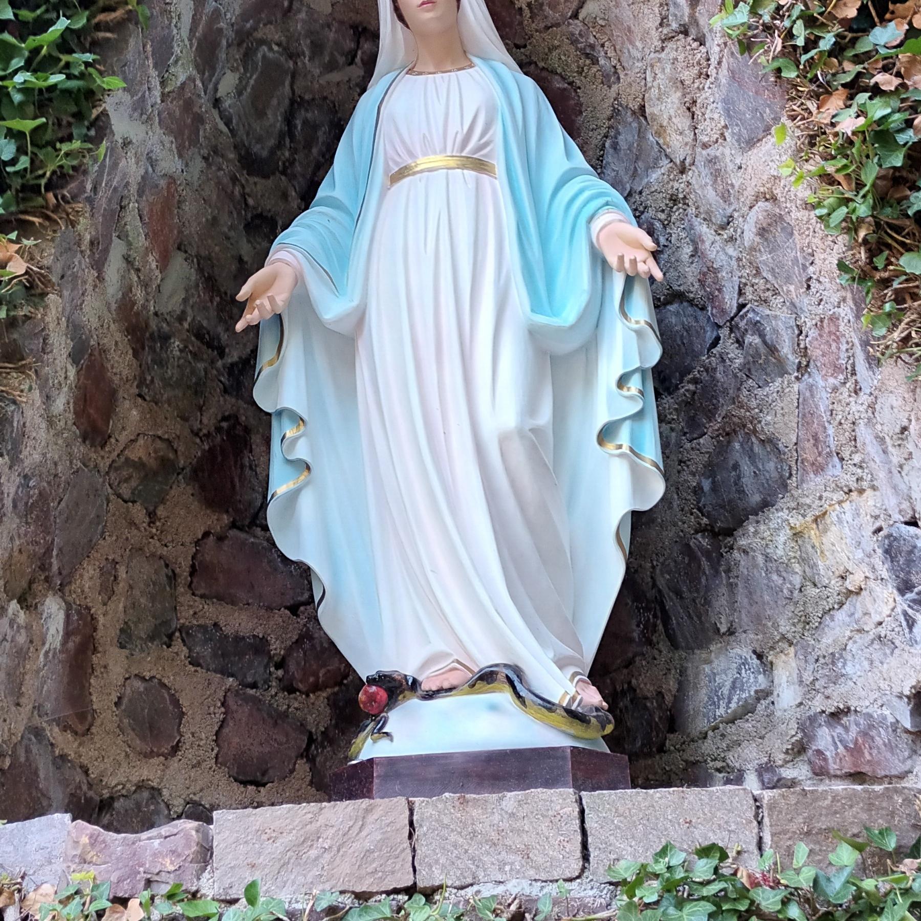 Statue der Jungfrau Maria in einer steinernen Nische, umgeben von Efeu. Maria trägt ein weißes Kleid mit goldenem Gürtel und einen blauen Mantel. Ihre Arme sind geöffnet, und zu ihren Füßen liegt eine Schlange mit einem roten Apfel