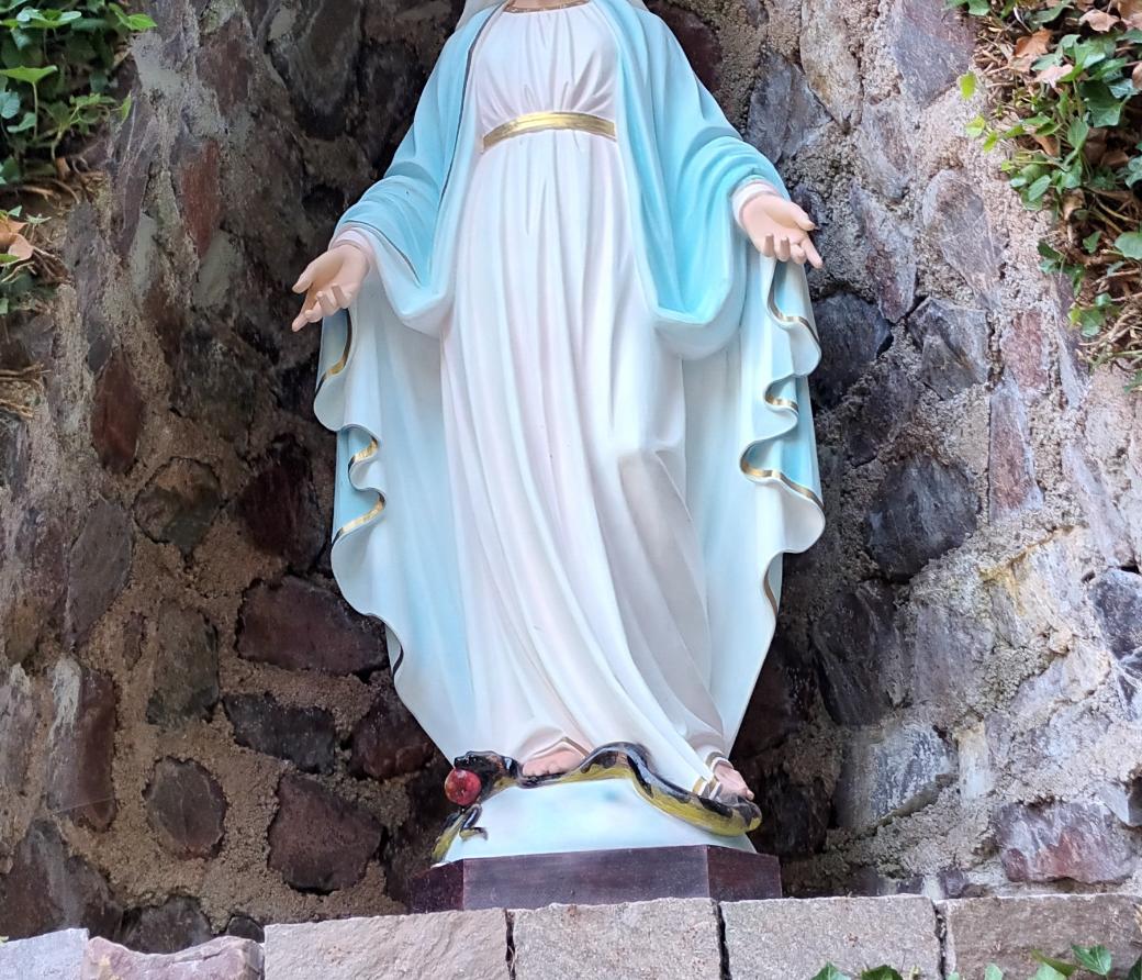 Statue der Jungfrau Maria in einer steinernen Nische, umgeben von Efeu. Maria trägt ein weißes Kleid mit goldenem Gürtel und einen blauen Mantel. Ihre Arme sind geöffnet, und zu ihren Füßen liegt eine Schlange mit einem roten Apfel