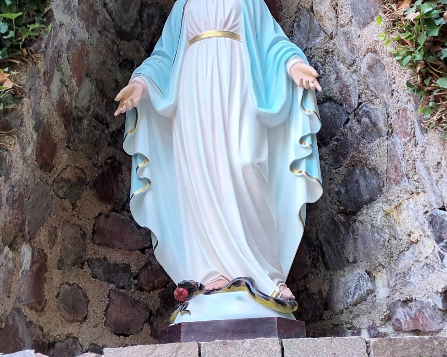 Statue der Jungfrau Maria in einer steinernen Nische, umgeben von Efeu. Maria trägt ein weißes Kleid mit goldenem Gürtel und einen blauen Mantel. Ihre Arme sind geöffnet, und zu ihren Füßen liegt eine Schlange mit einem roten Apfel