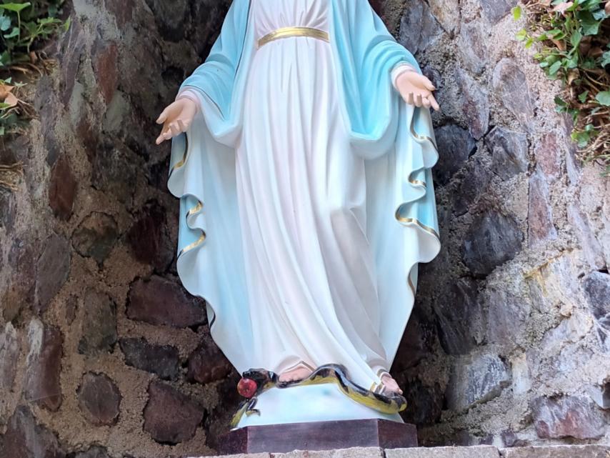 Statue der Jungfrau Maria in einer steinernen Nische, umgeben von Efeu. Maria trägt ein weißes Kleid mit goldenem Gürtel und einen blauen Mantel. Ihre Arme sind geöffnet, und zu ihren Füßen liegt eine Schlange mit einem roten Apfel