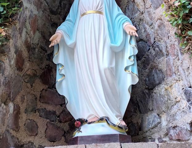 Statue der Jungfrau Maria in einer steinernen Nische, umgeben von Efeu. Maria trägt ein weißes Kleid mit goldenem Gürtel und einen blauen Mantel. Ihre Arme sind geöffnet, und zu ihren Füßen liegt eine Schlange mit einem roten Apfel