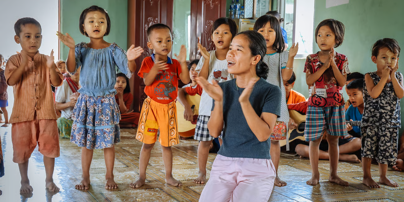 Eine Frau kniet auf einem gefliesten Boden und klatscht gemeinsam mit einer Gruppe junger Kinder, die in einer Reihe stehen. Die Kinder singen oder sprechen fröhlich mit und tragen bunte Kleidung. Im Hintergrund sitzen weitere Kinder und Erwachsene in einem hellgrünen Raum mit offenen Fenstern. Die Szene wirkt lebendig und gemeinschaftlich. (c) missio - glauben.leben.geben Eine Frau kniet auf einem gefliesten Boden und klatscht gemeinsam mit einer Gruppe junger Kinder, die in einer Reihe stehen. Die Kinder singen oder sprechen fröhlich mit und tragen bunte Kleidung. Im Hintergrund sitzen weitere Kinder und Erwachsene in einem hellgrünen Raum mit offenen Fenstern. Die Szene wirkt lebendig und gemeinschaftlich.