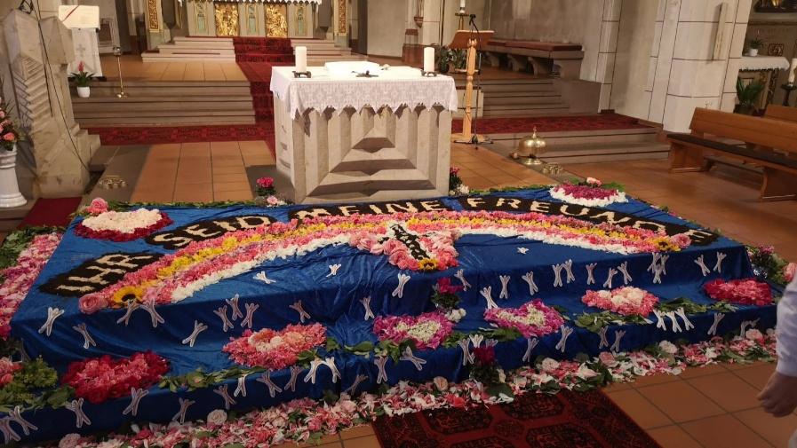 Foto eines kunstvoll gestalteten Blumenteppichs zu Fronleichnam in einer Kirche. Vor dem Altar ist ein großes blaues Tuch mit vielen Blüten, Symbolen und einem farbigen Kreuz ausgelegt. Auf dem Blumenteppich sind unter anderem Blumenmuster und religiöse Gestaltungselemente zu sehen. Im Hintergrund stehen der Altar, Kerzen und der Kirchenraum. Die Szene wirkt festlich, feierlich und liebevoll vorbereitet. (c) Pfarrei Saarwellingen St. Josef Foto eines kunstvoll gestalteten Blumenteppichs zu Fronleichnam in einer Kirche. Vor dem Altar ist ein großes blaues Tuch mit vielen Blüten, Symbolen und einem farbigen Kreuz ausgelegt. Auf dem Blumenteppich sind unter anderem Blumenmuster und religiöse Gestaltungselemente zu sehen. Im Hintergrund stehen der Altar, Kerzen und der Kirchenraum. Die Szene wirkt festlich, feierlich und liebevoll vorbereitet.