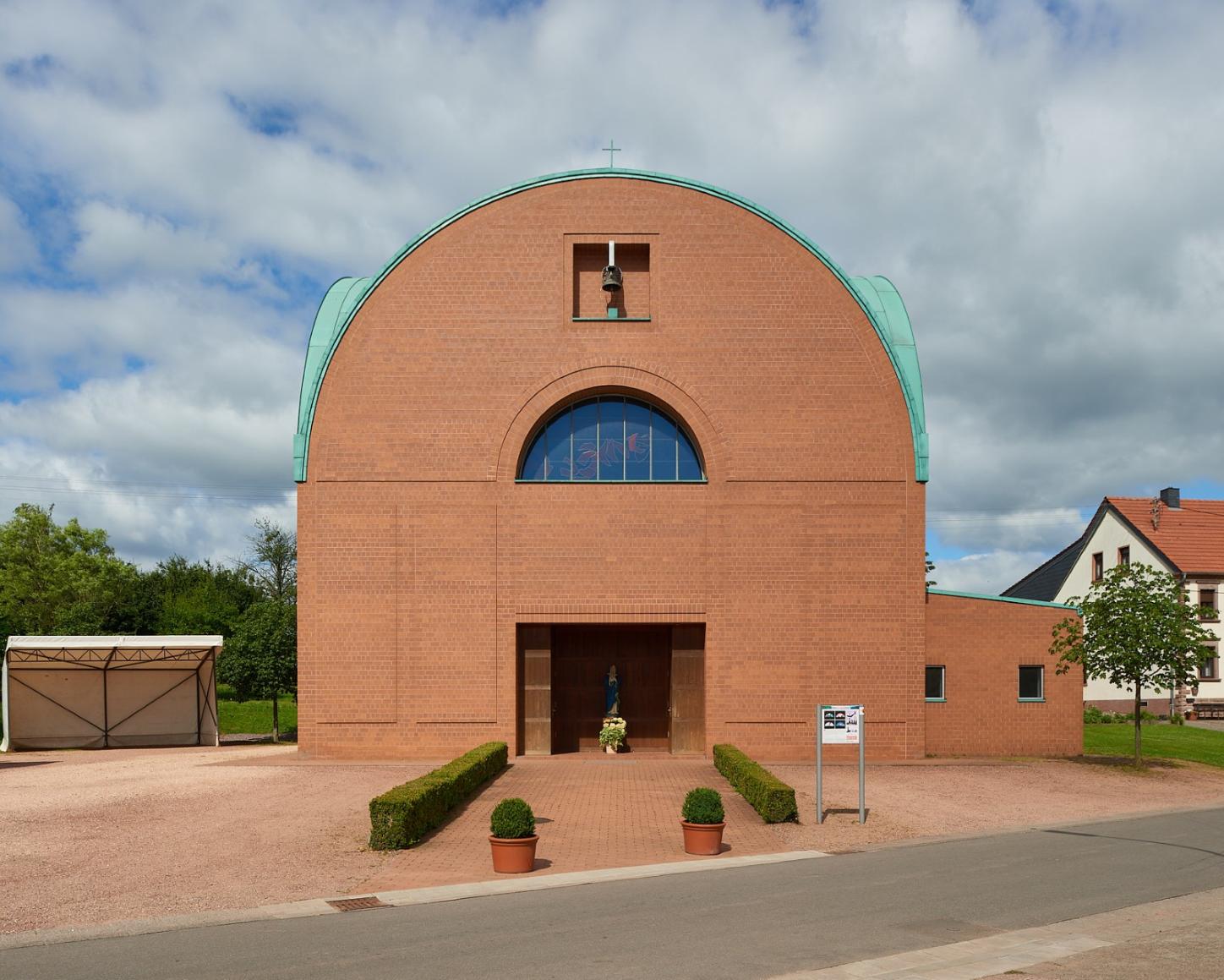 Backsteinkirche mit halbkreisförmigem Dachabschluss und grünem Kupferdach. Über dem Eingang befindet sich ein kleines Glockenfenster sowie ein rundbogiges Fenster mit buntem Glas. Vor dem Eingang steht eine Marienstatue mit Blumen. Links neben der Kirche ein Carport, rechts Wohnhäuser mit roten Dächern, im Vordergrund eine kleine Straße