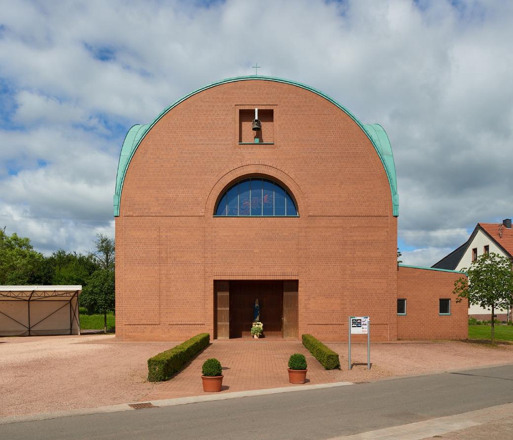 Backsteinkirche mit halbkreisförmigem Dachabschluss und grünem Kupferdach. Über dem Eingang befindet sich ein kleines Glockenfenster sowie ein rundbogiges Fenster mit buntem Glas. Vor dem Eingang steht eine Marienstatue mit Blumen. Links neben der Kirche ein Carport, rechts Wohnhäuser mit roten Dächern, im Vordergrund eine kleine Straße