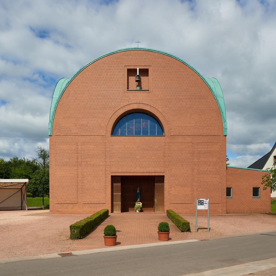 Backsteinkirche mit halbkreisförmigem Dachabschluss und grünem Kupferdach. Über dem Eingang befindet sich ein kleines Glockenfenster sowie ein rundbogiges Fenster mit buntem Glas. Vor dem Eingang steht eine Marienstatue mit Blumen. Links neben der Kirche ein Carport, rechts Wohnhäuser mit roten Dächern, im Vordergrund eine kleine Straße