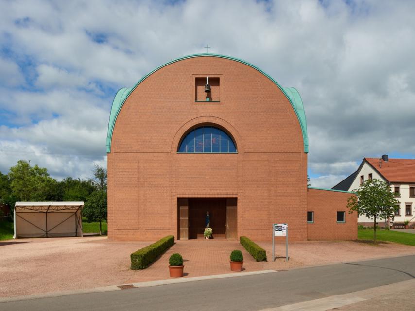 Backsteinkirche mit halbkreisförmigem Dachabschluss und grünem Kupferdach. Über dem Eingang befindet sich ein kleines Glockenfenster sowie ein rundbogiges Fenster mit buntem Glas. Vor dem Eingang steht eine Marienstatue mit Blumen. Links neben der Kirche ein Carport, rechts Wohnhäuser mit roten Dächern, im Vordergrund eine kleine Straße