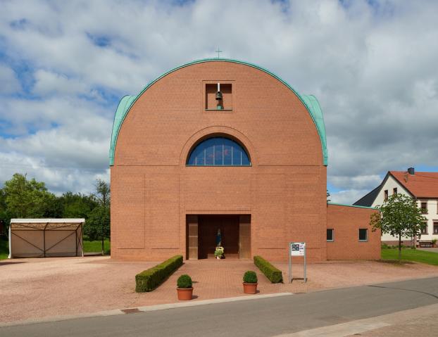 Backsteinkirche mit halbkreisförmigem Dachabschluss und grünem Kupferdach. Über dem Eingang befindet sich ein kleines Glockenfenster sowie ein rundbogiges Fenster mit buntem Glas. Vor dem Eingang steht eine Marienstatue mit Blumen. Links neben der Kirche ein Carport, rechts Wohnhäuser mit roten Dächern, im Vordergrund eine kleine Straße
