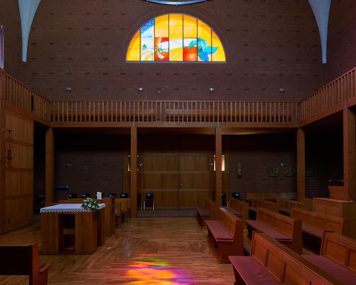 Innenraum einer modernen Backsteinkirche mit Holzvertäfelungen, Holzbänken und einem schlichten Altar. Über dem Eingang befindet sich ein großes rundbogiges Buntglasfenster mit abstrakten, farbenfrohen Motiven. Das einfallende Licht wirft bunte Reflexionen auf den Holzboden. An den Wänden sind Figuren und Kreuzwegdarstellungen angebracht