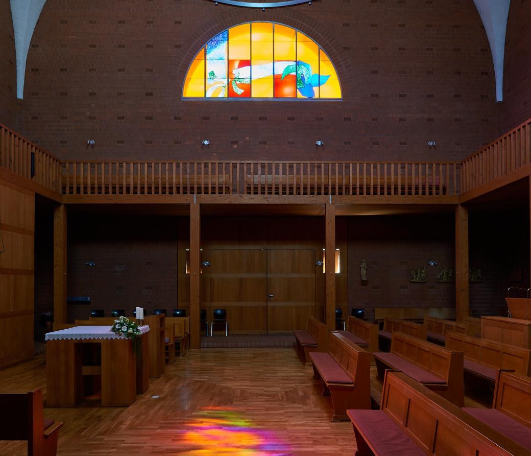 Innenraum einer modernen Backsteinkirche mit Holzvertäfelungen, Holzbänken und einem schlichten Altar. Über dem Eingang befindet sich ein großes rundbogiges Buntglasfenster mit abstrakten, farbenfrohen Motiven. Das einfallende Licht wirft bunte Reflexionen auf den Holzboden. An den Wänden sind Figuren und Kreuzwegdarstellungen angebracht