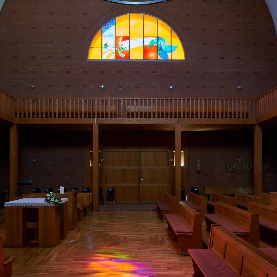 Innenraum einer modernen Backsteinkirche mit Holzvertäfelungen, Holzbänken und einem schlichten Altar. Über dem Eingang befindet sich ein großes rundbogiges Buntglasfenster mit abstrakten, farbenfrohen Motiven. Das einfallende Licht wirft bunte Reflexionen auf den Holzboden. An den Wänden sind Figuren und Kreuzwegdarstellungen angebracht