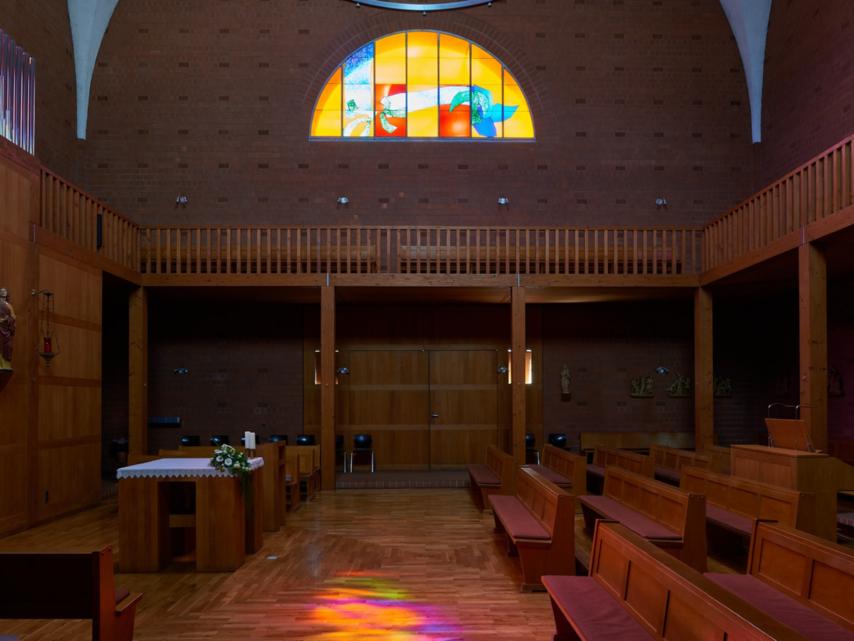 Innenraum einer modernen Backsteinkirche mit Holzvertäfelungen, Holzbänken und einem schlichten Altar. Über dem Eingang befindet sich ein großes rundbogiges Buntglasfenster mit abstrakten, farbenfrohen Motiven. Das einfallende Licht wirft bunte Reflexionen auf den Holzboden. An den Wänden sind Figuren und Kreuzwegdarstellungen angebracht
