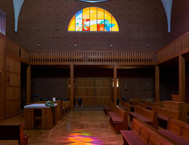Innenraum einer modernen Backsteinkirche mit Holzvertäfelungen, Holzbänken und einem schlichten Altar. Über dem Eingang befindet sich ein großes rundbogiges Buntglasfenster mit abstrakten, farbenfrohen Motiven. Das einfallende Licht wirft bunte Reflexionen auf den Holzboden. An den Wänden sind Figuren und Kreuzwegdarstellungen angebracht