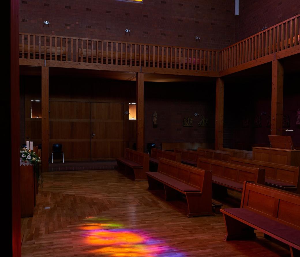 Innenraum einer modernen Kirche mit Holzvertäfelung und Holzbänken. Ein großes buntes Rundbogenfenster strahlt in kräftigen Farben und wirft farbige Lichtreflexionen in Gelb, Orange, Pink, Blau und Grün auf den Holzboden des Mittelgangs