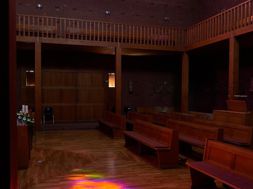 Innenraum einer modernen Kirche mit Holzvertäfelung und Holzbänken. Ein großes buntes Rundbogenfenster strahlt in kräftigen Farben und wirft farbige Lichtreflexionen in Gelb, Orange, Pink, Blau und Grün auf den Holzboden des Mittelgangs