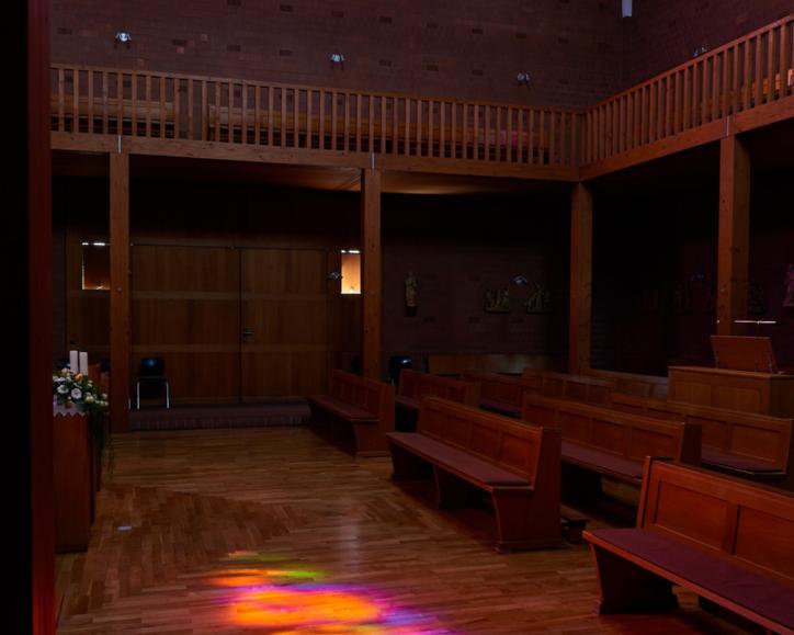 Innenraum einer modernen Kirche mit Holzvertäfelung und Holzbänken. Ein großes buntes Rundbogenfenster strahlt in kräftigen Farben und wirft farbige Lichtreflexionen in Gelb, Orange, Pink, Blau und Grün auf den Holzboden des Mittelgangs