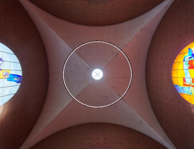 Blick nach oben auf die Decke einer Kirche: In der Mitte ein rundes Oberlicht mit Lichtkranz, seitlich zwei farbige Rundbogenfenster. Links zeigt ein blau-gelbes Glasfenster abstrakte Vogelmotive, rechts ein orangefarbenes Fenster mit blauen und grünen Formen
