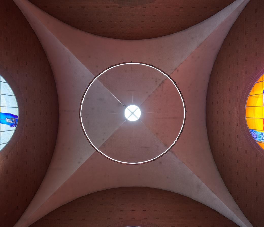 Blick nach oben auf die Decke einer Kirche: In der Mitte ein rundes Oberlicht mit Lichtkranz, seitlich zwei farbige Rundbogenfenster. Links zeigt ein blau-gelbes Glasfenster abstrakte Vogelmotive, rechts ein orangefarbenes Fenster mit blauen und grünen Formen