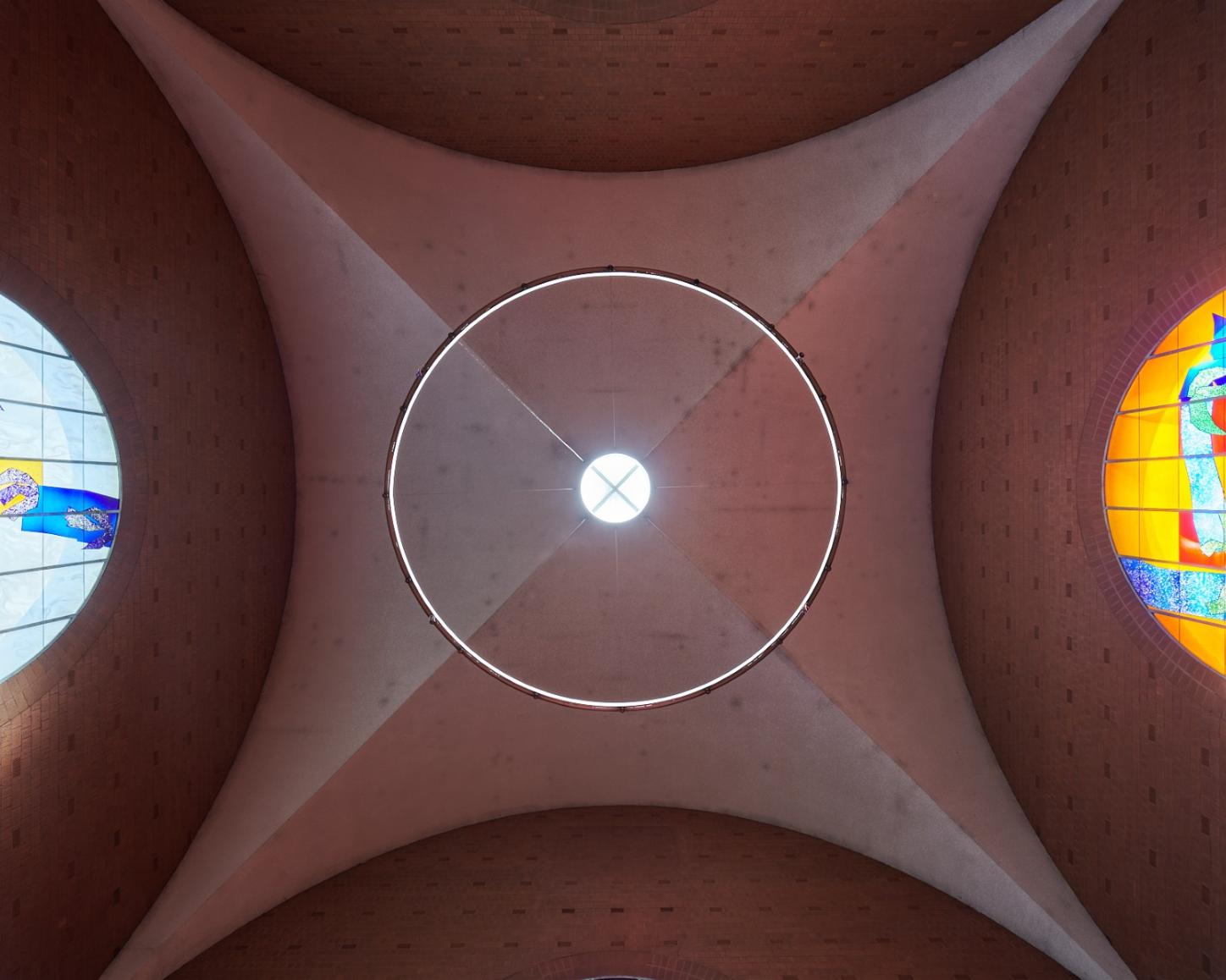 Blick nach oben auf die Decke einer Kirche: In der Mitte ein rundes Oberlicht mit Lichtkranz, seitlich zwei farbige Rundbogenfenster. Links zeigt ein blau-gelbes Glasfenster abstrakte Vogelmotive, rechts ein orangefarbenes Fenster mit blauen und grünen Formen