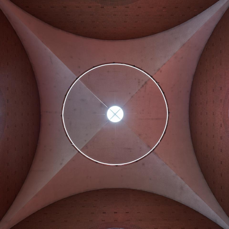 Blick nach oben auf die Decke einer Kirche: In der Mitte ein rundes Oberlicht mit Lichtkranz, seitlich zwei farbige Rundbogenfenster. Links zeigt ein blau-gelbes Glasfenster abstrakte Vogelmotive, rechts ein orangefarbenes Fenster mit blauen und grünen Formen