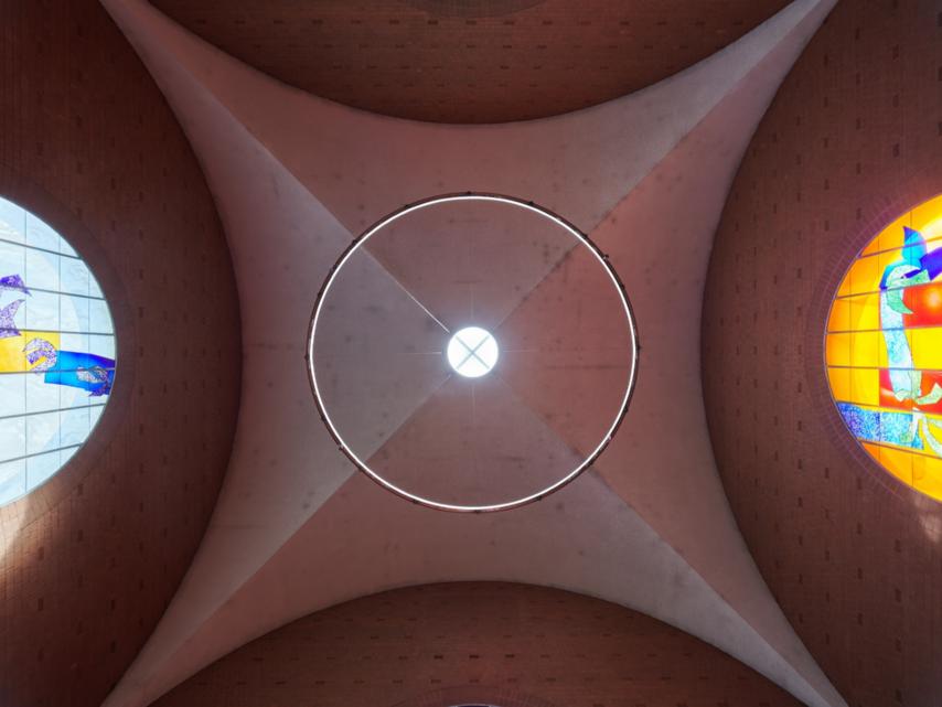 Blick nach oben auf die Decke einer Kirche: In der Mitte ein rundes Oberlicht mit Lichtkranz, seitlich zwei farbige Rundbogenfenster. Links zeigt ein blau-gelbes Glasfenster abstrakte Vogelmotive, rechts ein orangefarbenes Fenster mit blauen und grünen Formen