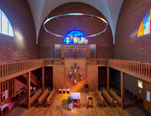 Innenraum einer Kirche mit Blick auf den Altarraum. In der Mitte ein schlichter Altar mit weißem Tuch, darüber ein großes Holzkruzifix mit Christusfigur und zwei Heiligenstatuen. Dahinter eine Empore mit Orgelpfeifen und einem leuchtenden blauen Rundbogenfenster. An den Seiten zwei weitere bunte Glasfenster, links in Blau, rechts in Orange und Gelb. Über dem Raum hängt ein großer runder Lichtkranz.