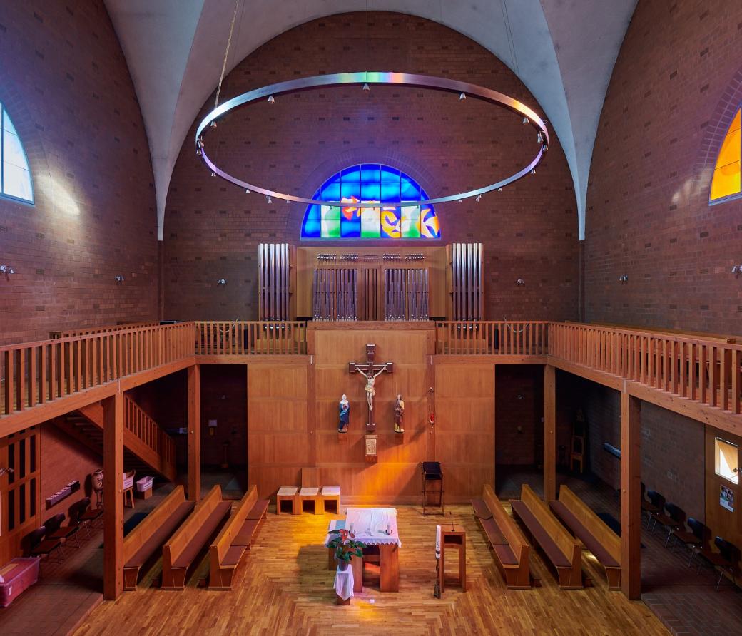 Innenraum einer Kirche mit Blick auf den Altarraum. In der Mitte ein schlichter Altar mit weißem Tuch, darüber ein großes Holzkruzifix mit Christusfigur und zwei Heiligenstatuen. Dahinter eine Empore mit Orgelpfeifen und einem leuchtenden blauen Rundbogenfenster. An den Seiten zwei weitere bunte Glasfenster, links in Blau, rechts in Orange und Gelb. Über dem Raum hängt ein großer runder Lichtkranz.
