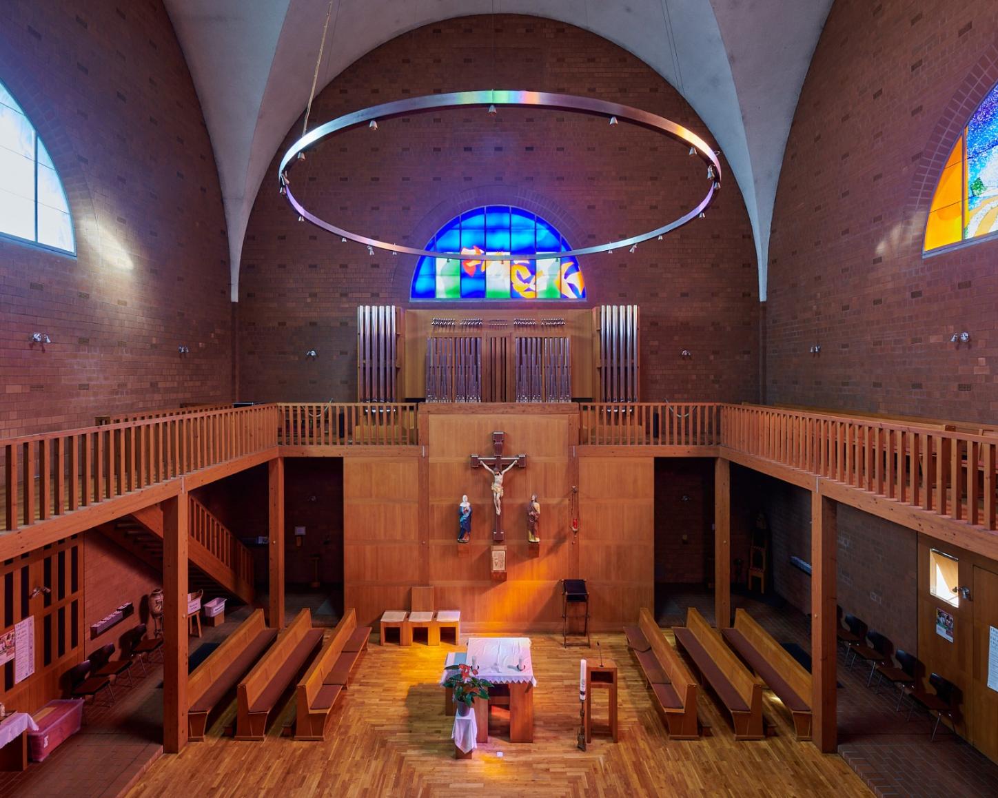 Innenraum einer Kirche mit Blick auf den Altarraum. In der Mitte ein schlichter Altar mit weißem Tuch, darüber ein großes Holzkruzifix mit Christusfigur und zwei Heiligenstatuen. Dahinter eine Empore mit Orgelpfeifen und einem leuchtenden blauen Rundbogenfenster. An den Seiten zwei weitere bunte Glasfenster, links in Blau, rechts in Orange und Gelb. Über dem Raum hängt ein großer runder Lichtkranz.
