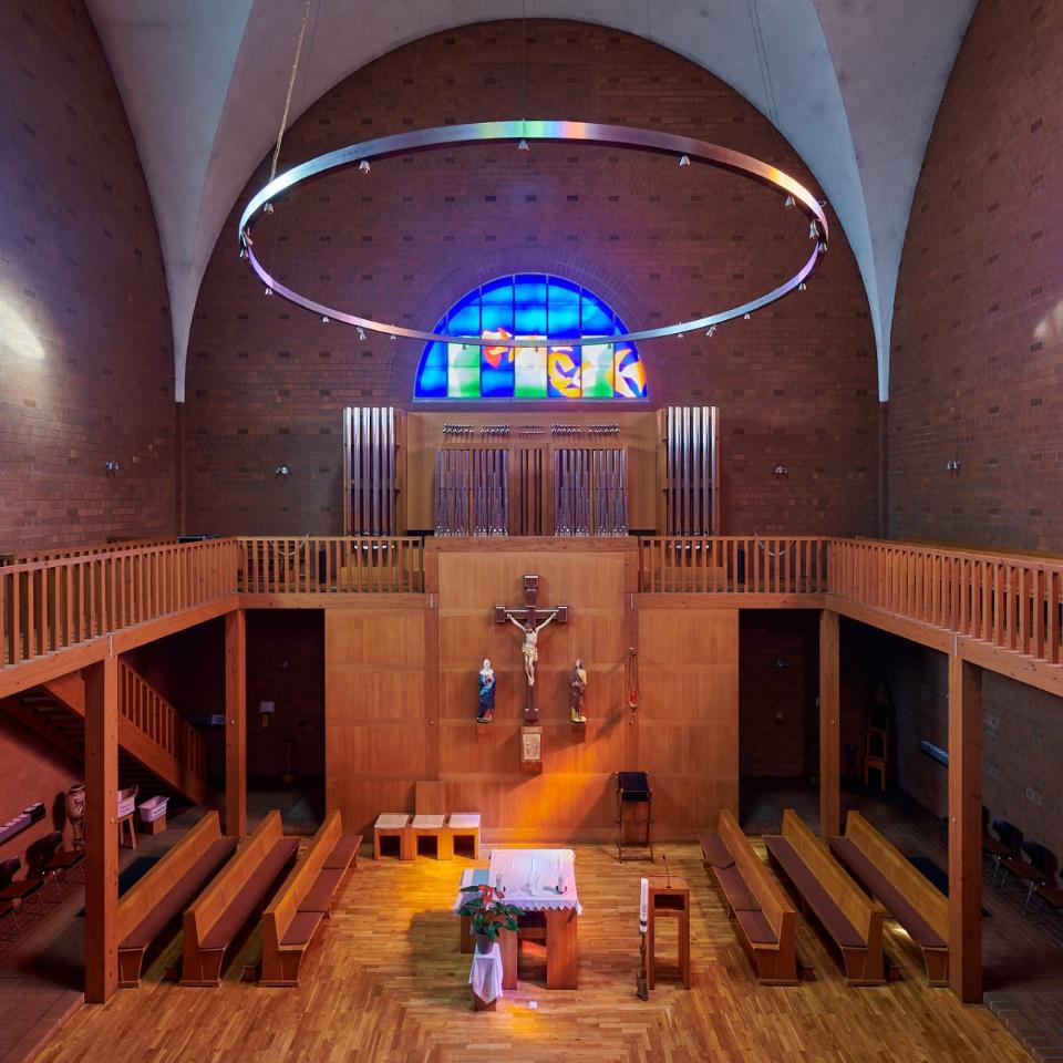 Innenraum einer Kirche mit Blick auf den Altarraum. In der Mitte ein schlichter Altar mit weißem Tuch, darüber ein großes Holzkruzifix mit Christusfigur und zwei Heiligenstatuen. Dahinter eine Empore mit Orgelpfeifen und einem leuchtenden blauen Rundbogenfenster. An den Seiten zwei weitere bunte Glasfenster, links in Blau, rechts in Orange und Gelb. Über dem Raum hängt ein großer runder Lichtkranz.