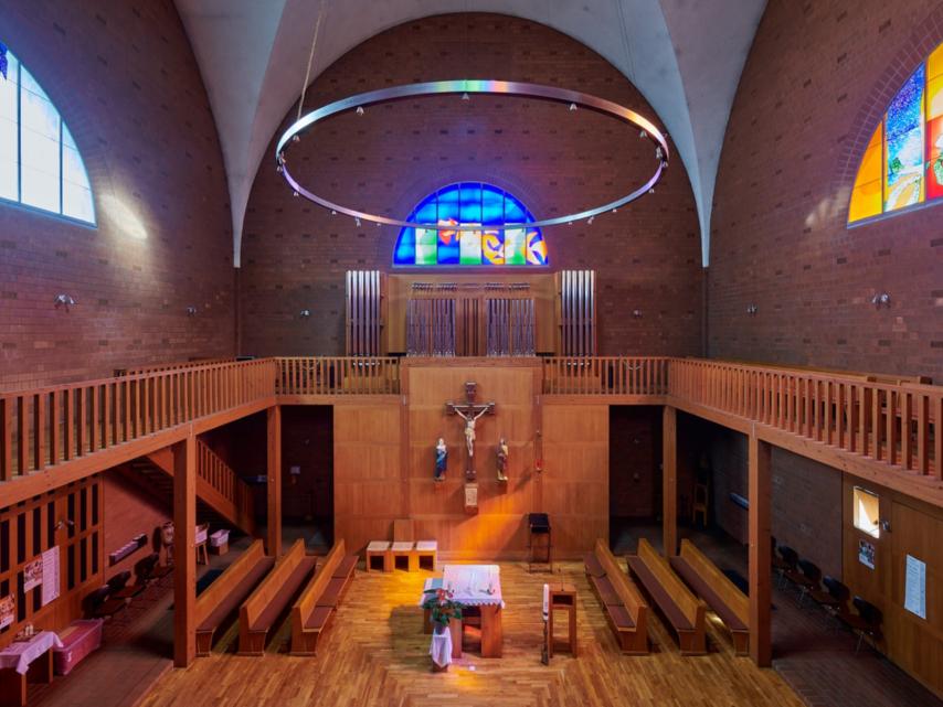 Innenraum einer Kirche mit Blick auf den Altarraum. In der Mitte ein schlichter Altar mit weißem Tuch, darüber ein großes Holzkruzifix mit Christusfigur und zwei Heiligenstatuen. Dahinter eine Empore mit Orgelpfeifen und einem leuchtenden blauen Rundbogenfenster. An den Seiten zwei weitere bunte Glasfenster, links in Blau, rechts in Orange und Gelb. Über dem Raum hängt ein großer runder Lichtkranz.