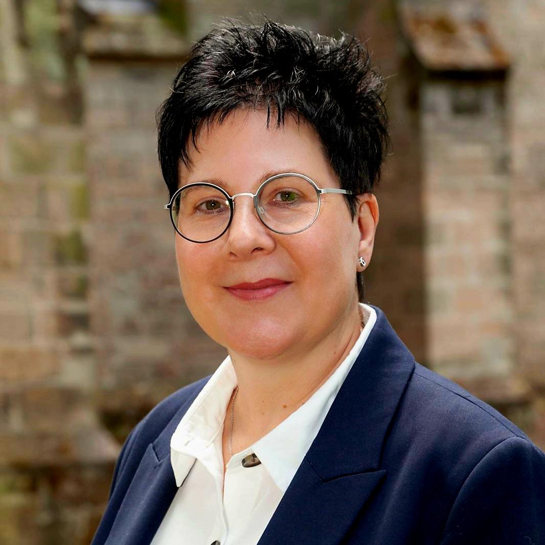Porträt von Stefanie Kallenborn. Sie trägt eine Brille, hat kurze dunkle Haare und ist in einem dunklen Blazer mit weißer Bluse vor der Pfarrkirche St. Blasius zu sehen