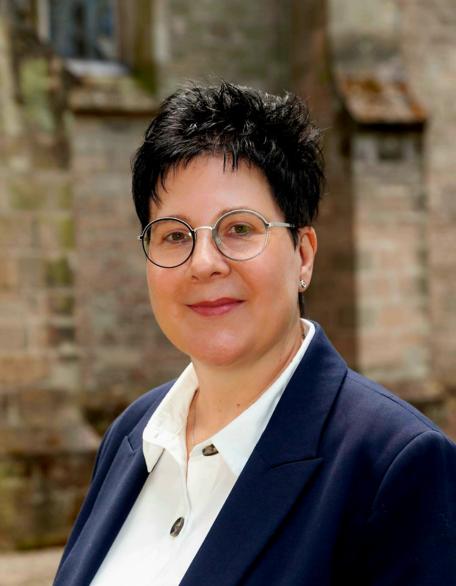 Porträt von Stefanie Kallenborn. Sie trägt eine Brille, hat kurze dunkle Haare und ist in einem dunklen Blazer mit weißer Bluse vor der Pfarrkirche St. Blasius zu sehen