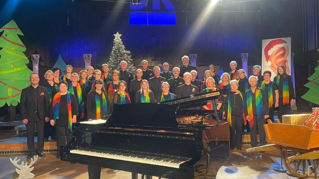 Auf einer Bühne steht ein gemischter Chor aus etwa 30 bis 40 Personen, überwiegend ältere Erwachsene, in mehreren Reihen. Alle tragen schwarze Kleidung mit bunten, regenbogenfarbenen Schals. Vor ihnen steht ein großer schwarzer Flügel. Im Hintergrund ist eine weihnachtliche Dekoration zu sehen, darunter ein geschmückter Weihnachtsbaum und stilisierte Tannenbäume. Die Bühne ist hell ausgeleuchtet, darüber hängt ein Schild mit der Aufschrift ‚SR‘. Die Szene wirkt wie ein festliches Konzert oder eine Aufführung zur Weihnachtszeit