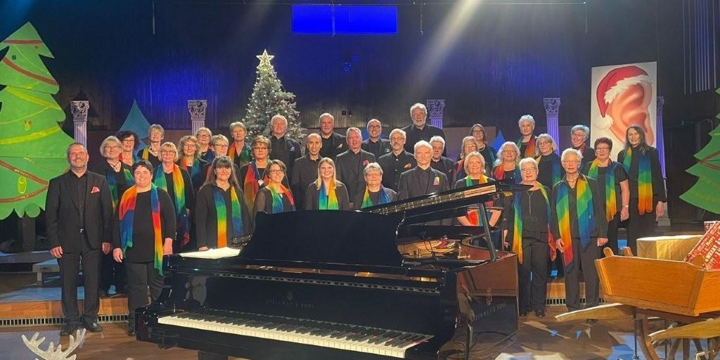 Auf einer Bühne steht ein gemischter Chor aus etwa 30 bis 40 Personen, überwiegend ältere Erwachsene, in mehreren Reihen. Alle tragen schwarze Kleidung mit bunten, regenbogenfarbenen Schals. Vor ihnen steht ein großer schwarzer Flügel. Im Hintergrund ist eine weihnachtliche Dekoration zu sehen, darunter ein geschmückter Weihnachtsbaum und stilisierte Tannenbäume. Die Bühne ist hell ausgeleuchtet, darüber hängt ein Schild mit der Aufschrift ‚SR‘. Die Szene wirkt wie ein festliches Konzert oder eine Aufführung zur Weihnachtszeit