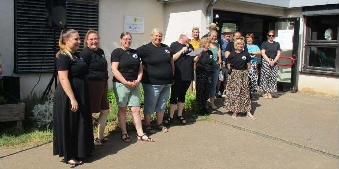 Foto einer Gruppe von Erwachsenen vor einem Gebäude an einem sonnigen Tag. Mehrere Personen stehen gemeinsam vor dem Eingang und wirken freundlich und zugewandt. Einige tragen schwarze Kleidung mit einem kleinen Logo, im Hintergrund sind Fenster, eine Tür und ein Schild am Gebäude zu sehen. Das Bild vermittelt Gemeinschaft, Begegnung und Engagement vor Ort. (c) Frau Saskia Müller Kita RB Foto einer Gruppe von Erwachsenen vor einem Gebäude an einem sonnigen Tag. Mehrere Personen stehen gemeinsam vor dem Eingang und wirken freundlich und zugewandt. Einige tragen schwarze Kleidung mit einem kleinen Logo, im Hintergrund sind Fenster, eine Tür und ein Schild am Gebäude zu sehen. Das Bild vermittelt Gemeinschaft, Begegnung und Engagement vor Ort.